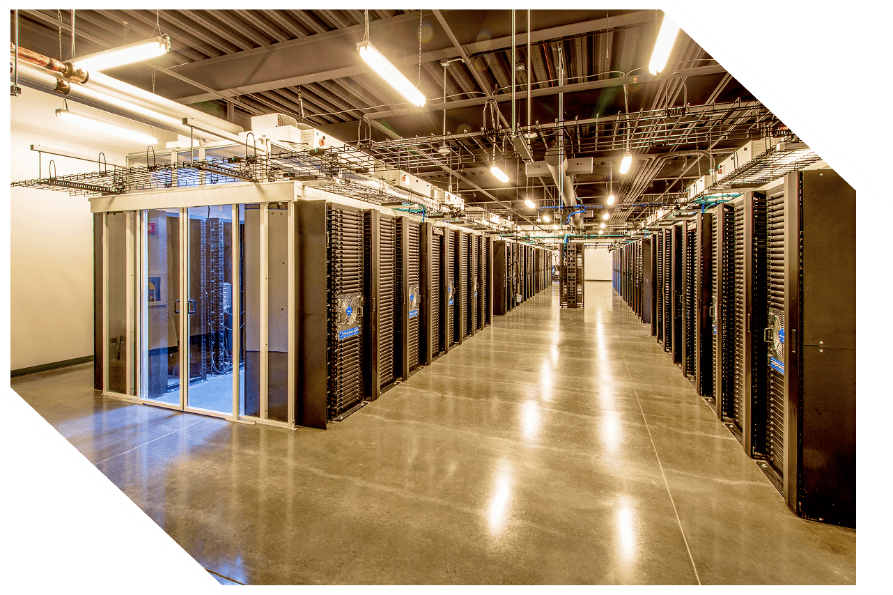 A brightly lit data center with rows of server racks. The hallway leads to a glass door at one end. Overhead cabling is visible above the racks.