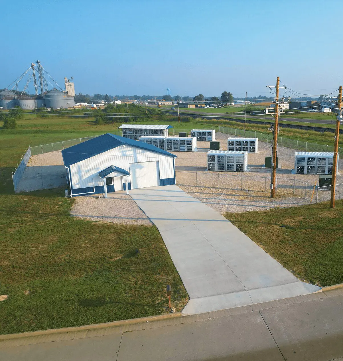 Small industrial facility with a white building, fenced area, and multiple outdoor units. Surrounded by green fields and nearby silos under a clear blue sky.