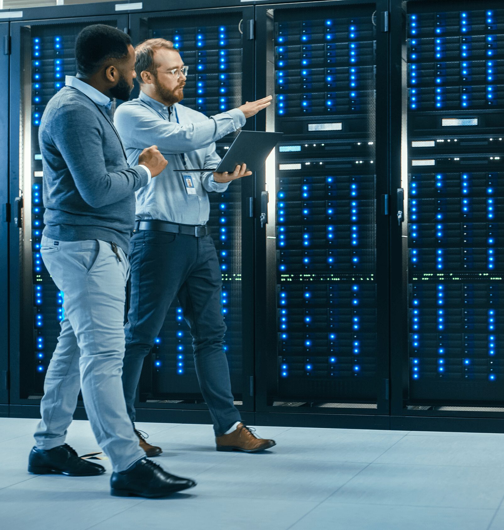 Two men stand in a server room, discussing while looking at a tablet. They are surrounded by racks of computer servers with blue indicator lights.