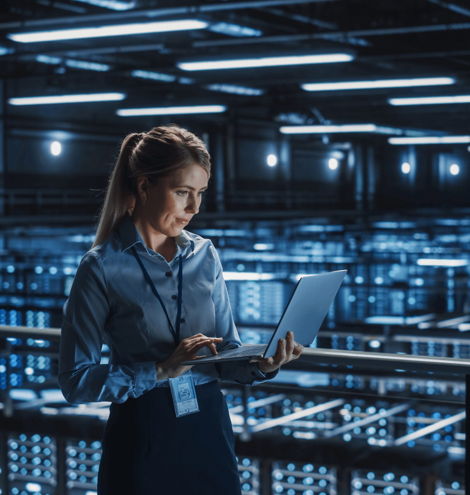 A person in a blue shirt stands in a data center, holding a laptop. Rows of server racks with blue lights are visible in the background.