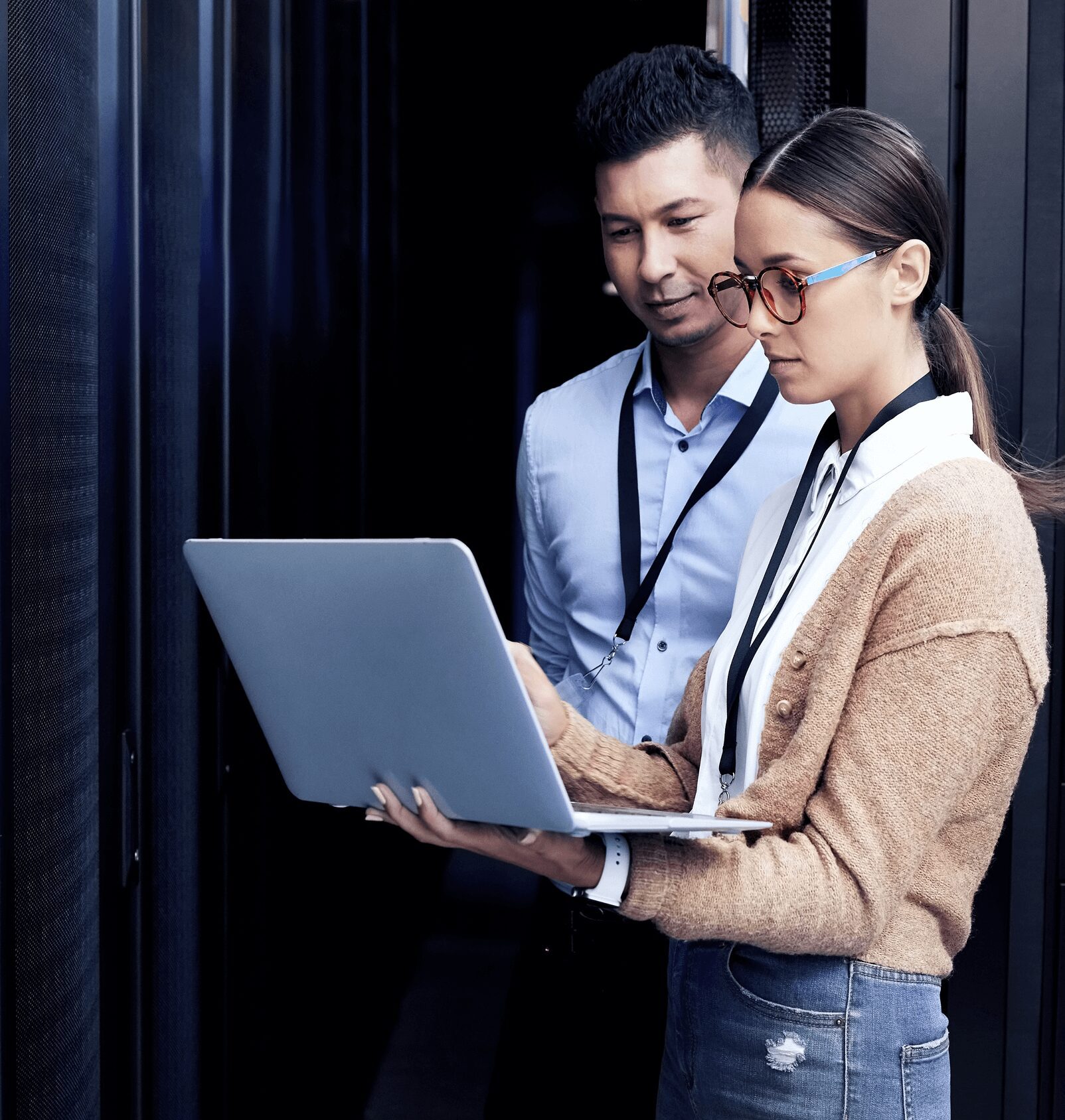 Two people in a server room examining a laptop screen. They both wear lanyards and appear focused on the task.