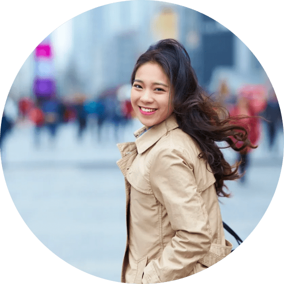 Smiling person with long hair wearing a beige coat, standing outdoors in an urban area with a blurred background of people and buildings.