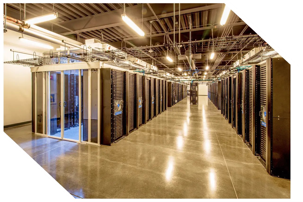 A brightly lit data center with rows of server racks. The hallway leads to a glass door at one end. Overhead cabling is visible above the racks.